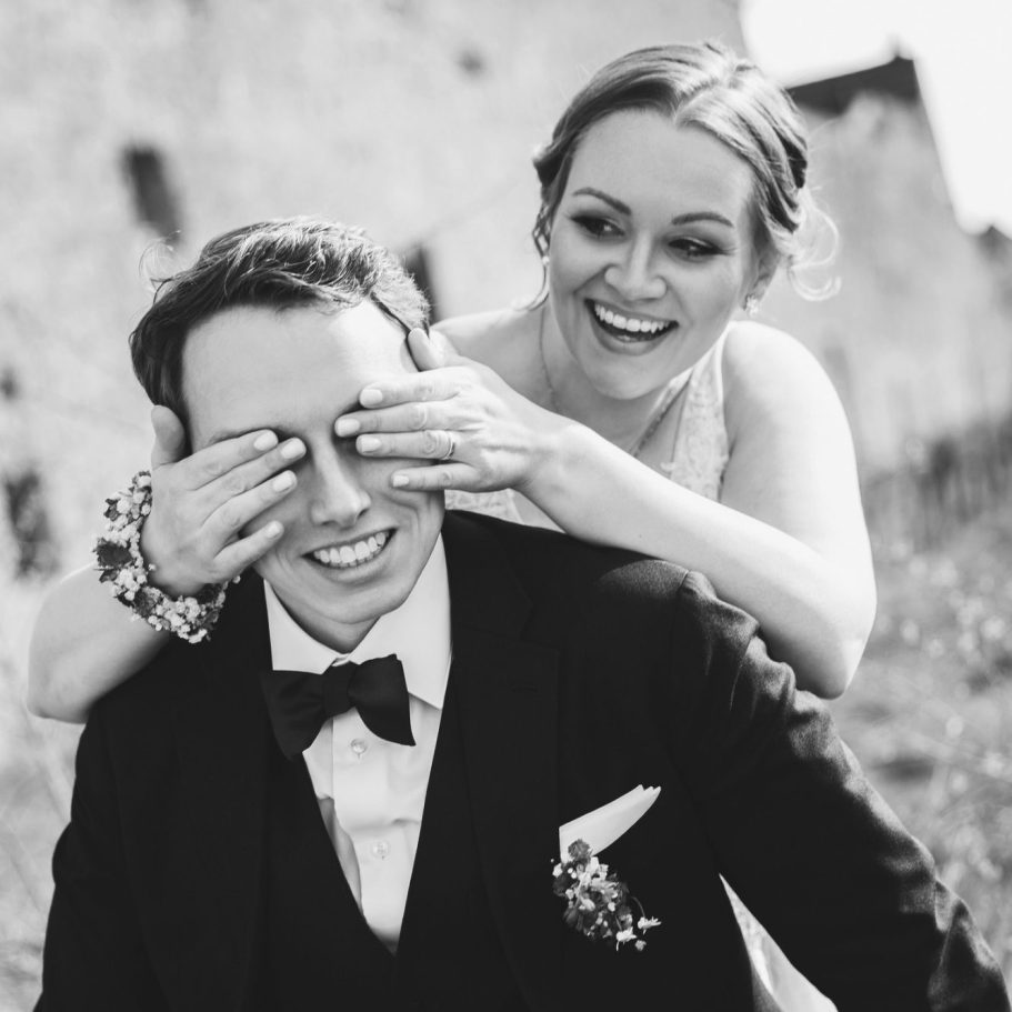 Braut verdeckt Bräutigam die Augen Braut hält dem sitzenden Bräutigam spielerisch die Augen zu, fotografiert in Schwarzweiß.