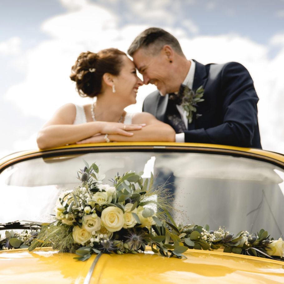 Brautpaar auf Oldtimer Motorhaube Hochzeitspaar lehnt sich lachend auf dekorierten Oldtimer.