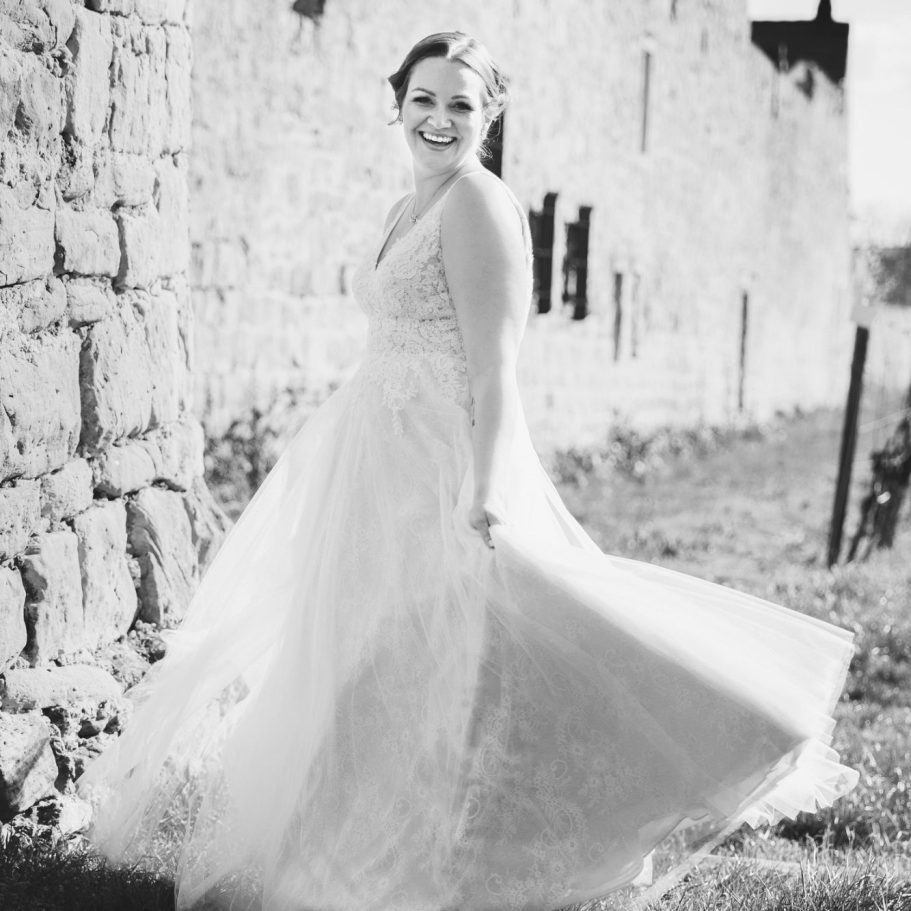 Braut im Brautkleid vor Steinmauer Braut steht lachend im Brautkleid vor einer alten Steinmauer, fotografiert in Schwarzweiß.