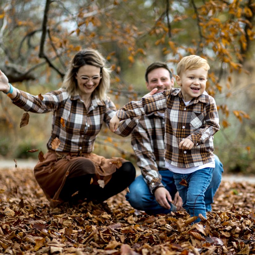 Familienfoto mit Kind im Herbst Kind läuft lachend durch Herbstlaub, Eltern folgen im Hintergrund bei natürlichem Licht.