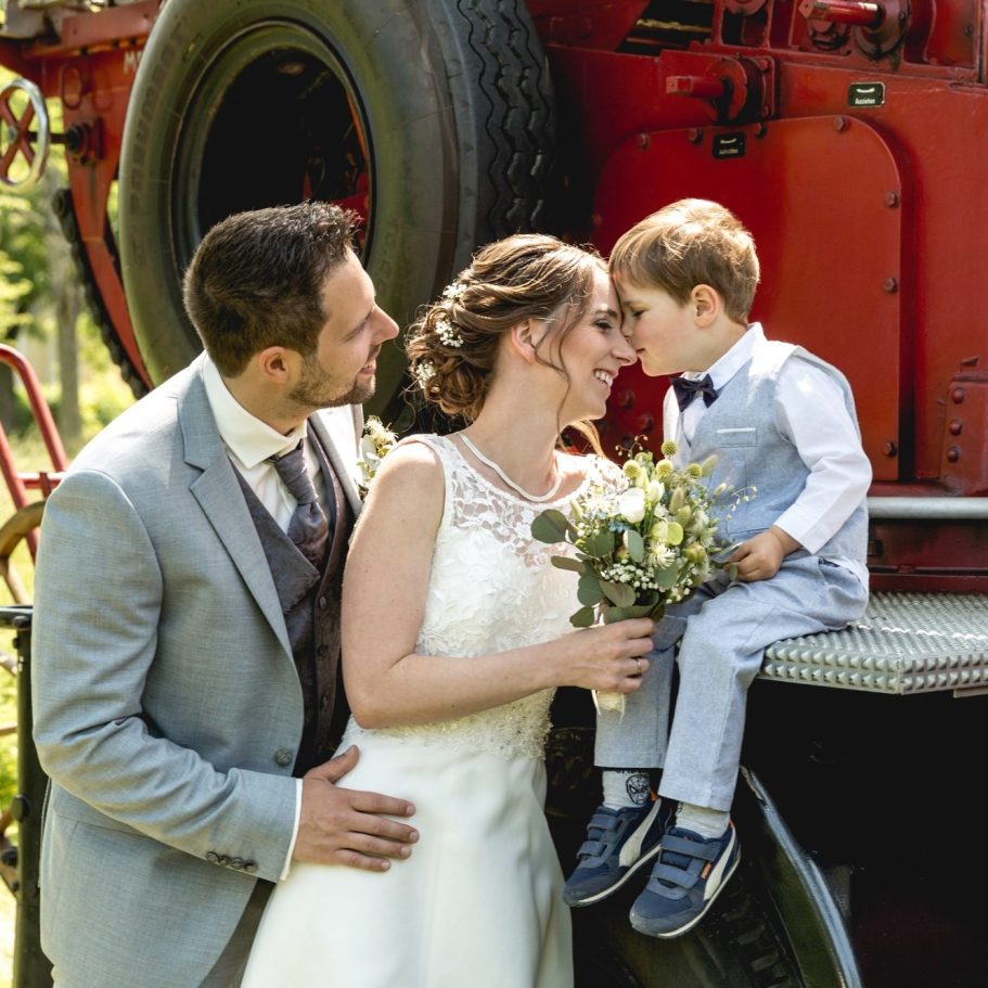 Brautpaar mit Kind vor einem Oldtimer der Feuerwehr Brautpaar mit kleinem Jungen posiert vor einem roten historischen Oldtimer im Freien.