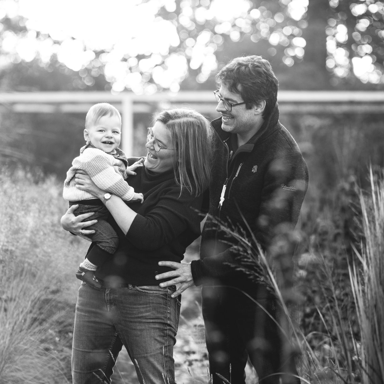 Schwarzweiß Portrait einer Familie mit Baby Familie mit Baby steht zwischen hohen Gräsern bei natürlichem Licht, fotografiert in Schwarzweiß.