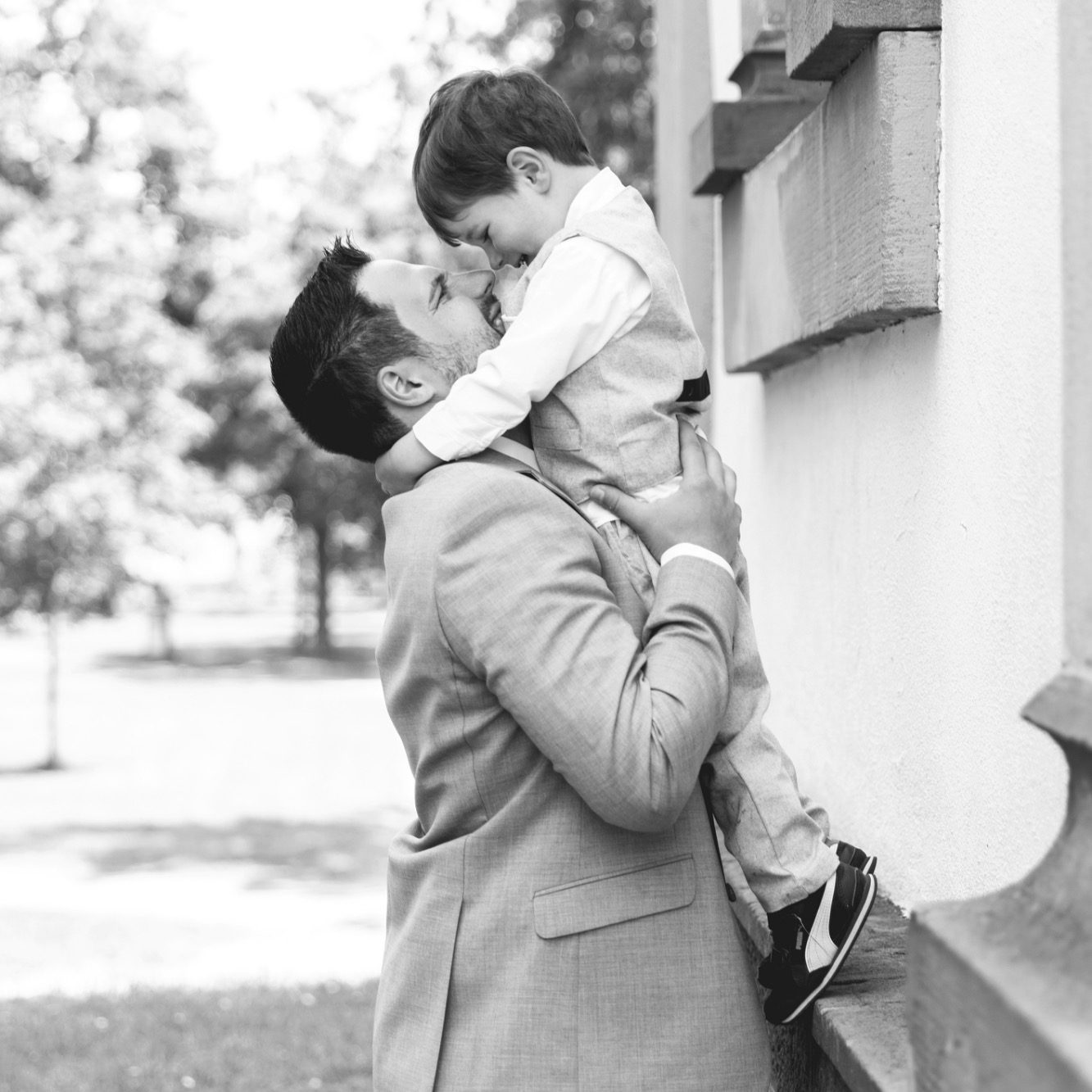 Vater hebt Kind hoch bei Hochzeit Mann im Anzug hebt ein Kind an einer Hauswand hoch, fotografiert in Schwarzweiß.