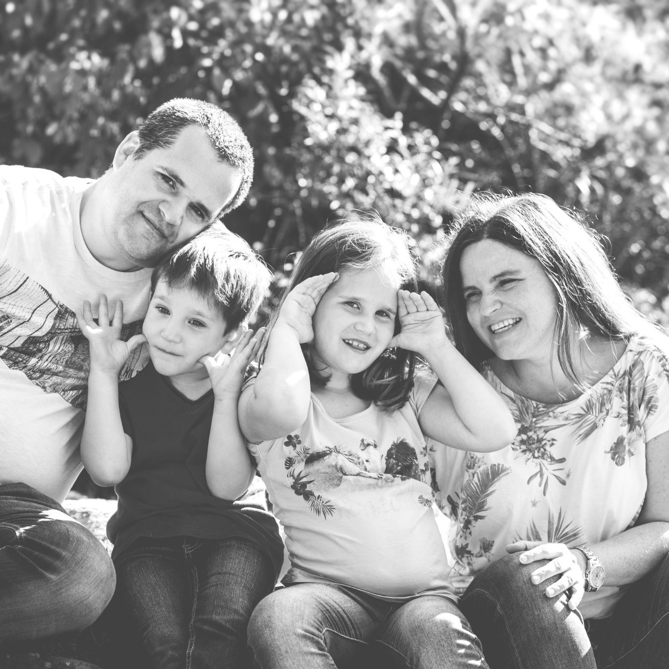 Schwarzweiß Familienportrait mit zwei Kindern Eltern sitzen mit zwei Kindern im Freien und machen gemeinsam eine spielerische Geste.