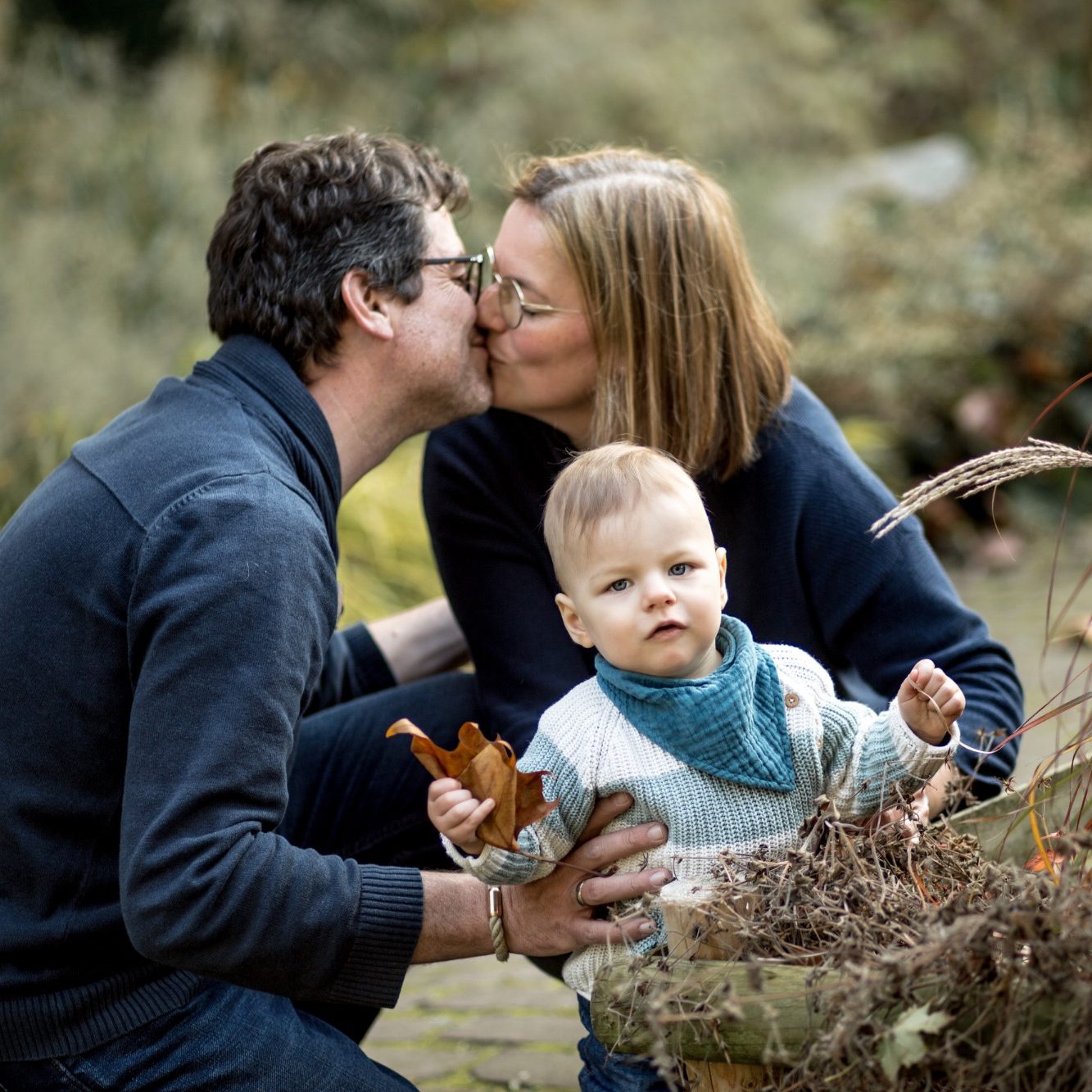 Eltern küssen sich mit Baby im Vordergrund Baby im Vordergrund, Eltern küssen sich im Hintergrund bei einem natürlichen Outdoor-Fotoshooting.